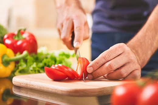 concepto de cocina, comida y hogar: primer plano de una mano masculina cortando un tomate en una tabla de cortar en casa
