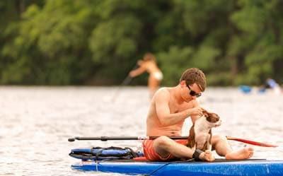 Man and Boston terrier sitting on a blue paddle board in the middle of a lake.